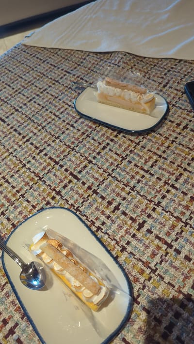 A rectangular piece of cake sits on a white oval plate with a blue rim. The cake has a yellow base, a white cream topping, and is garnished with toasted meringue and slivered almonds. A silver spoon rests to the left of the cake on the plate. A second, similar piece of cake is visible on another white oval plate in the upper right portion of the frame. Both plates are set on a textured, multicolored fabric surface. The setting appears to be indoors. The location is Larkollen, Norway.