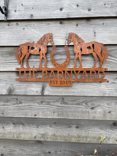 The image depicts a rusty metal sign affixed to a weathered, grey wooden fence. The sign features two horses facing opposite directions, a horseshoe in the center, and the name "RINARD" vertically aligned along the right side, followed by "EST 2019" underneath. The fence is constructed of vertical planks with visible wood grain, knots, and nail heads. A hint of greenery, possibly a branch, is visible on the right side of the fence. A metal gate mechanism is partially visible in the lower-left corner. The scene's location is not discernible from the image.