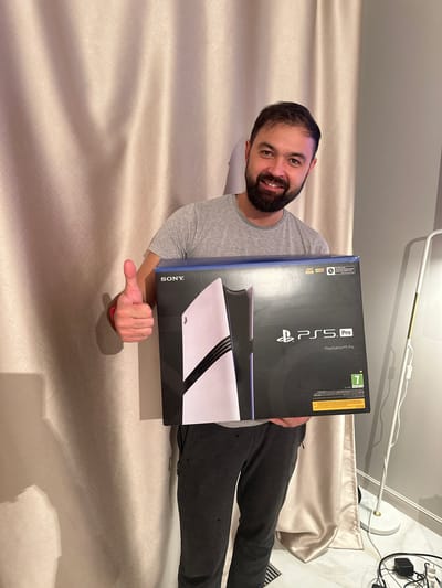 A man is depicted in an indoor setting, likely in Edem, Russia, lying or reclining against a light-colored, subtly patterned fabric background. He is smiling broadly at the camera, holding a black box for a PlayStation 5 Pro console. His right hand is visible, giving a thumbs-up gesture, conveying excitement or satisfaction.
The man has a beard and short dark hair, and he is wearing a grey t-shirt and dark sweatpants. The PlayStation 5 Pro box prominently features the console's design in black and white. Visible text on the box includes "PS5 Pro," "PlayStation®5 Pro," "SONY," and technical indicators such as "4K," "HDR," "120 FPS," and "8K Output." A green age rating symbol with the number "7" is also clearly seen.
In the background, white cables, a light fixture, and a power adapter are partially visible, suggesting a typical indoor environment. The man's joyful expression and the brand-new console box indicate a moment of happiness over a recent acquisition or gift. The lighting appears to be artificial.
