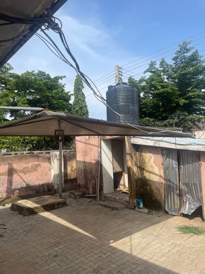 This media file captures an outdoor scene in Kporva, Nigeria. The image is oriented vertically, presenting a view looking upwards. In the background, a large black water tank is visible, mounted on a pole alongside numerous power lines crisscrossing against a clear blue sky with scattered white clouds. Lush green trees frame the scene on the left and in the distance.

In the foreground, a weathered building with peeling pink and yellow paint dominates the right side of the frame. The roof of a structure, possibly a lean-to or awning, extends from the building. The ground is paved with interlocking concrete blocks, and a distinct shadow suggests it is daytime, likely mid-morning or afternoon, with the sun positioned to the right and slightly behind the viewer.

The building appears to be aged, with signs of disrepair, including corrugated metal sheeting on the upper part of its exterior and what looks like barbed wire near the roofline. There are open doorways and partially visible interior spaces. A bright blue plastic container or jug is placed on the ground near a section of damp, discolored wall. The overall impression is of a utilitarian, possibly residential or commercial, area in an African setting. There are no people visible in the frame, and no text is discernible.