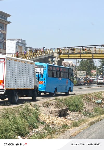 A bustling street scene features a white truck and a blue minibus labeled "SIMBA" parked side by side. Above them, a yellow pedestrian bridge is crowded with people. A row of vehicles lines the adjacent road, leading towards distant trees and buildings under a bright, clear sky. In the foreground, a drainage ditch is lined with vegetation and litter. The scene conveys a sense of everyday urban life in a developing area.
