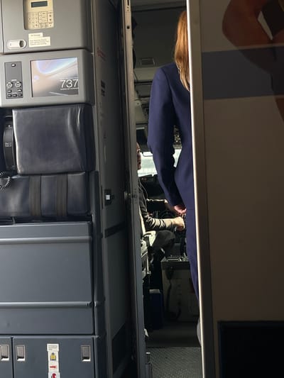 The rotated image, viewed upright, depicts the interior of an aircraft, specifically looking from the galley or forward cabin towards an open cockpit.
In the foreground, a person with long blonde hair, wearing a dark blue uniform jacket, stands at the threshold of the cockpit, observed from the back and side. Inside the cockpit, another individual, also in a uniform, is seated and appears to be interacting with the flight controls or other cockpit equipment. Faintly visible above the cabin features, a third person's face can be seen, possibly lying in a crew rest bunk.
The cabin area in the lower part of the frame features various control panels and storage units. On the left, there is a panel with a numerical keypad. Below it, a warning sign reads, "No Stowage" and "Door Must Be Closed and Latched During Taxi, Takeoff, and Landing." Adjacent to this, a screen prominently displays "NEXT-GENERATION 737 BOEING SKY INTERIOR" and "737," indicating the aircraft type. Below this screen is another control interface with buttons and a small display showing "00." To the right, several dark grey padded sections, possibly jump seats or equipment covers, are secured with straps. Further right, a series of grey cabinets or drawers are visible, one of which carries a label "Emergency Use Only" alongside icons for a flashlight and an oxygen mask. A coiled cable, likely for a crew interphone, is also present.
The scene suggests a period of pre-flight checks or preparations, with crew members active around the cockpit. The lighting is moderate, with some areas appearing dimmer than others. The overall setting is a modern aircraft interior. This media file was captured in Quart de Poblet, Spain.