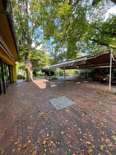 This image captures an unoccupied outdoor patio area in La Wantzenau, France, under daylight conditions, exhibiting clear signs of autumn. The ground consists primarily of reddish-brown rectangular paver bricks, heavily scattered with dry, yellowish-brown deciduous leaves. A distinct gray square concrete patch is embedded within the brickwork in the mid-ground.
To the left, a building presents a bright yellow facade with multiple large, dark-framed glass windows or doors. A white rain gutter system is visible along the roofline of this building. Immediately adjacent to the building stands a mature tree with a thick, textured trunk, its canopy displaying a mix of green and yellowing foliage.
The right and center portions of the scene are dominated by an extensive outdoor structure featuring a light brown fabric awning, supported by multiple slender white metal poles. This awning provides overhead cover for an arrangement of dark-colored tables and chairs, suggesting an outdoor dining or lounge area. Several wooden structures, possibly benches or planters, are also visible within this covered space. Further in the background, beyond the covered area, additional outdoor seating elements are discernible on a grassy surface.
The environment is characterized by mature trees with ample green and yellowing leaves, casting dappled shadows across the patio. The sky, visible through breaks in the canopy, is clear and blue. No individuals are present in the scene.