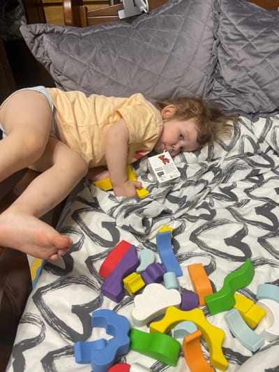 A young child wearing a peach-colored shirt and white underwear lies on a bed with a white and black patterned comforter. The bed is covered in a gray pillow. The child is holding a small rectangular box with an image of a brown cartoon animal. The child's right foot is on the bed, and the left foot is dangling off the edge. The child is looking down at a set of wooden toys with a variety of shapes and colors. The wooden toys are scattered on the bed around the child. The coordinates provided are for the city of Moscow, Russia. This image appears to be taken in a private residence.