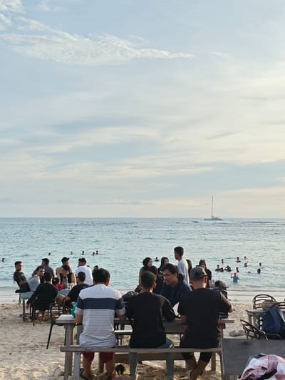 The image depicts a beach scene in Monikeun, Indonesia. A group of people are seated at picnic tables on the sand, seemingly enjoying a meal or gathering. In the background, people are swimming and wading in the ocean. A sailboat is visible on the horizon. The sky is partly cloudy.