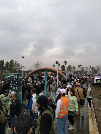 A crowd gathers under a cloudy sky, some holding flags of Syria. Police cars with flashing lights are visible in the background. An arch of green and orange balloons marks a starting or ending point. Individuals on horseback mingle within the group. Many people wear helmets. The ground appears wet and muddy. Trees and buildings are visible in the distance, along with palm trees.