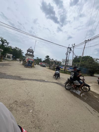 The image depicts an outdoor scene in Rantau Pauh, Indonesia, on an overcast day. The foreground is dominated by a rough, unpaved road with visible patches of water and gravel. In the middle ground, two individuals are riding a black motorcycle, with one person driving and another passenger. They are wearing casual clothing. To the left of the road, a dark colored motorcycle with a sidecar is parked. In the background, there is a large gate structure with a tower, which appears to be an entrance to a facility. Signs are visible on the gate, including one that reads "PT. PERTAMINA" and another with "OBJEK VITAL NASIONAL". A white building and trees are also present behind the gate. Further back, more trees and a cloudy sky are visible. Electrical poles and wires are prominent throughout the image, indicating infrastructure. To the right of the road, another motorcycle is visible from the side, and a light-colored vehicle is parked further down the road. The overall environment appears to be an industrial or administrative area.
