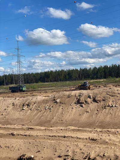 The image shows a construction site in Luzhitsy, Russia. A dirt pit or excavation area is visible on the right side of the frame. A bulldozer is positioned along the edge of a treeline. Further down, a green truck with a trailer is situated beside an electricity pylon supporting high-voltage power lines. Orange marker balls are attached to the power lines. The sky is blue with scattered white clouds.