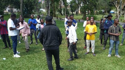 A group of around 20 young men stand on a grassy lawn surrounded by trees and buildings. Most of the men are looking in the same direction, while one writes on a clipboard. A man in a black jacket faces away from the camera, seemingly addressing the group. The men's clothing varies, with casual shirts, jeans, and sneakers being the most common. Some wear hats or caps. The setting appears to be a park or a campus.