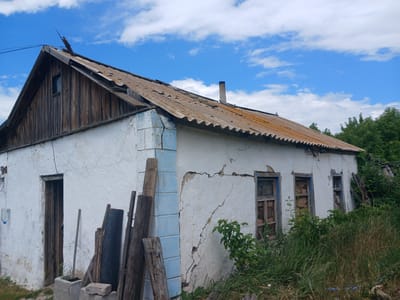 A dilapidated, white stucco house stands under a bright blue sky with scattered white clouds in the foreground of the image, located in the city of Zhaltyr, Kazakhstan. The house has a dilapidated, corrugated metal roof, weathered and discolored with age. The walls are cracked and peeling in several places, revealing the underlying material. A wooden gable end on the left side of the house has vertical wooden planks and a small, dark window. To the right of the gable, the roof slopes down, with a brick chimney visible near the peak. The house has several windows, some with broken glass and others boarded up. In front of the house, there is overgrown grass and weeds, and several pieces of wood are leaning against the wall near the entrance. A plastic water bottle is hanging from a wire on the left side of the house, near an open doorway. The overall scene depicts a neglected and abandoned property.