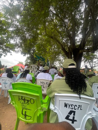 The image depicts an outdoor gathering of individuals, primarily young adults, seated on plastic chairs under the shade of a large tree. The attendees are dressed in matching green shirts and khaki shorts, consistent with the uniform of the National Youth Service Corps (NYSC) in Nigeria. Many are wearing green caps. The chairs are a mix of white and bright green plastic. Some white chairs have "NYSC-PL" and a number stenciled on them. The ground appears to be dirt or sand. In the background, there are glimpses of more people, some red-roofed structures, and dense foliage. One person in the foreground is partially visible, showing their back and braided hair. The setting suggests an NYSC orientation camp or a similar official gathering. The presence of "Emure-Ekiti" on some of the chairs indicates the location is likely Emure-Ekiti, Nigeria.