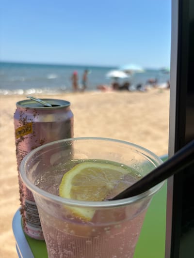 The image captures a close-up view of a pink can of Schweppes and a clear plastic cup filled with a pink beverage, ice, and a lemon slice. A black straw is immersed in the drink. The can and cup are placed on a green surface, which appears to be a table. In the background, a blurred beach scene unfolds, with the sea, sand, and several people enjoying the sunny weather. Beach umbrellas are visible in the distance. The location is identified as Osenovo, Bulgaria.