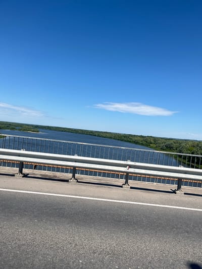 The image shows a view from a bridge over a wide river. The sky is clear and blue with a few white clouds. The river is a deep blue color and is bordered by lush green trees on both sides. A metal guardrail runs along the side of the bridge, with an asphalt road in the foreground. The location is identified as the city of Yadrin, Russia. There are no people or animals visible in the image. The scene is tranquil and showcases a natural landscape.