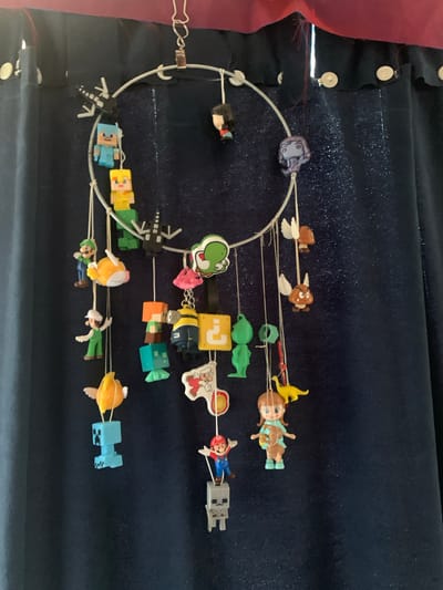 This is a medium shot, eye-level, indoor photograph of a mobile hanging in front of a dark blue, glittery curtain. The mobile is circular and made of white wire, with various small figurines suspended from it by thin white strings. The figurines include characters from video games such as Minecraft (Steve, a Creeper, Alex, a skeleton), Super Mario (Luigi, a Goomba, Mario), and a Minion. There are also other objects like a yellow question mark block, a yellow dinosaur, and a stylized orange fish. The mobile appears to be a DIY craft project. The lighting is natural and comes from an unseen source to the left, casting subtle shadows. The overall impression is one of a playful and whimsical decoration. The location of Сонячне, Ukraine is not discernible from the image itself.