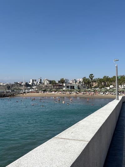 The image depicts a sunny day at a beach in Chorrillos, Peru. In the foreground, a concrete barrier runs along the edge of a body of water, likely the Pacific Ocean, which is a deep blue-green. The water is populated by numerous individuals swimming and wading. In the midground, a sandy beach is visible, lined with palm trees and umbrellas providing shade for beachgoers. Buildings, including apartment complexes and commercial structures, are situated on a hill overlooking the beach. A streetlamp stands on the right side of the image, illuminating the scene. A small airplane is visible in the upper left corner of the sky, and a faint crescent moon can be seen in the upper right.