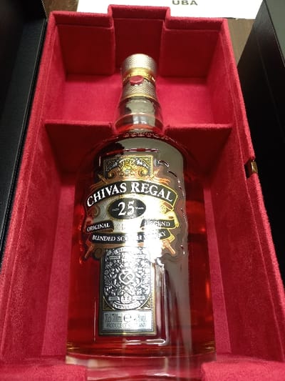 A bottle of Chivas Regal 25 Year Old Blended Scotch Whisky is prominently displayed within an open, red velvet-lined presentation box. The clear glass bottle contains an amber-colored liquid and features a gold-toned, ribbed screw cap with a small red circular seal. The main label reads "CHIVAS REGAL 25 Years ORIGINAL BLENDED SCOTCH WHISKY," with a lower label detailing "70cl / 700ml e" and "40% vol.," indicating production in Scotland. The interior of the box is molded to cradle the bottle, while its exterior, partially visible, is black. A white object displaying the letters "UBA" is visible in the upper background. The scene is located in the city of Oturkpo, Nigeria.