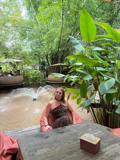 A woman is sitting on a large, orange beanbag chair next to a wooden table. She is wearing a leopard-print, one-shoulder dress and a necklace. In front of her is a shallow pool of water with a small fountain. The area appears to be an outdoor seating area for a restaurant or cafe, with more seating and umbrellas visible in the background. Lush green vegetation, including bamboo and large-leafed plants, surrounds the scene. The image was taken in Tambon Mae Raem, Thailand.