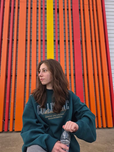 A young female is sitting in front of a colorful striped wall in Chita, Russia. The wall is made of vertical slats painted in alternating shades of red, orange, and yellow. The woman is wearing a dark teal sweatshirt with white lettering and gray sweatpants. She has long, brown hair and is looking to her left. In her right hand, she is holding a clear plastic water bottle, with her thumb on the cap. The ground beneath her is a concrete or asphalt surface. The overall scene appears to be outdoors, possibly in an urban setting.