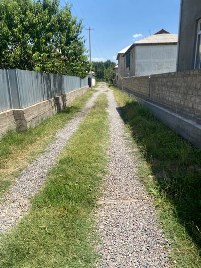 The image shows a narrow, unpaved path or driveway made of gravel, bordered by grass. On the left side, there is a tall, corrugated metal fence with a concrete foundation. On the right side, there is a low brick wall and the side of a building. The path recedes into the distance, where trees and more buildings are visible under a clear blue sky. The scene appears to be in a residential area with a rustic feel. The location is identified as Боғистон, Tajikistan.