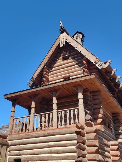 The image shows a detailed, close-up view of a traditional log cabin, characteristic of Russian architecture, against a clear blue sky. The cabin is constructed from dark brown, rough-hewn logs with visible chinking between them. It features a steeply pitched roof with decorative wooden carvings along the eaves, including what appear to be stylized bird figures and intricate patterns. A prominent covered porch with wooden railings and sturdy support posts extends from the upper level of the cabin. The log construction is evident throughout the visible portion of the building, with interlocking corner joinery clearly visible. The bright sunlight casts shadows, highlighting the texture of the wood and the details of the craftsmanship. No people are visible in the image. The specific location is identified as Терехово-2 Снт, Russia.