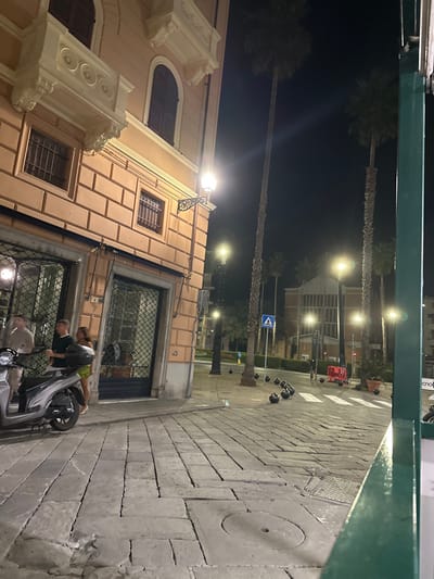 The image depicts a nighttime street scene in Sestri Levante, Italy, specifically looking towards the Piazza della Stazione with the Sestri Levante train station in the background.
In the foreground, a light orange, multi-story building with decorative white balconies and arched windows occupies the left side. A classic-style streetlamp extends from its corner. Below, on the cobbled pavement, a silver-grey scooter is parked. Three individuals are visible near the ground-floor entrance of the building: two males, one in a light shirt and one in a dark shirt, and a female partially visible in a light-colored skirt or shorts. They appear to be standing on the sidewalk.
The foreground pavement is composed of large, irregular grey cobblestones, with a circular metal manhole cover visible on the right. Beyond the building, a wide street or plaza is illuminated by multiple bright streetlights. Several tall palm trees are spaced along this area. Numerous black spherical bollards are scattered across the ground, and a pedestrian crossing (zebra stripes) is visible. A blue pedestrian crossing sign is also present. In the mid-ground right, red temporary barriers and potted plants are visible. The Sestri Levante train station, a prominent building with tall, narrow window sections and a gabled roof, stands in the background. The sky is dark, indicating night.
