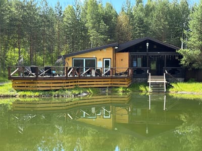 This image depicts a rustic wooden house situated on the edge of a calm, reflective body of water, likely a pond or lake. The house has a distinctive architectural style, with one section constructed from dark, horizontal logs and another from lighter, vertical wooden planks, creating a contrasting facade. A large, elevated wooden deck extends from the front of the house, furnished with lounge chairs and modern hanging chairs, suggesting an outdoor seating area. The deck is bordered by a wooden railing. A set of white stairs leads down from a darker section of the house to the water's edge. The background is dominated by a dense forest of tall, green pine trees. The water's surface mirrors the house, the deck, and the surrounding trees, creating a symmetrical effect. The scene appears to be set in a natural, possibly rural or semi-rural, environment. The context provided mentions Vorsma, Russia, suggesting this is the location of the property. There are no people visible in the image. The lighting indicates it is daytime, with clear skies and sunlight illuminating the scene.