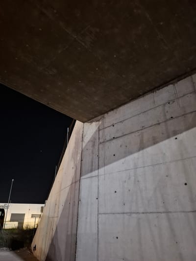 The image depicts the underside of a concrete overpass or bridge structure, likely at night. The location is identified as Quart de Poblet, Spain. Two concrete walls meet at an angle, framing a view of the night sky and a portion of a building and street. The concrete shows signs of wear and construction details such as bolt holes and seam lines. The lighting is dim, illuminating parts of the concrete structure and the nearby building. The building appears to be a low-rise commercial structure with windows and signage.