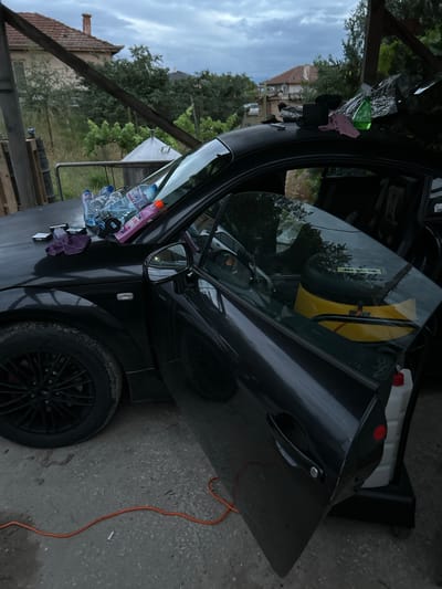 The image depicts a black Audi TT parked outdoors in Benkovski, Bulgaria, under a cloudy sky. The driver's side door is ajar, revealing the car's interior. On the hood of the car, several plastic bottles, a purple microfiber cloth, and packs of cigarettes are visible. A green spray bottle and a pink cloth are placed on the roof of the car. Inside the car, a yellow object, possibly a tool bag or a speaker, is visible. A white plastic container with a red cap is located next to the car, on the right side. An orange electrical cord lies on the ground in front of the car, with a cigarette butt near it. In the background, a house with a red tile roof and trees can be seen. The setting appears to be a yard or a garage area.
