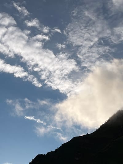 The image shows a silhouetted mountain range against a cloudy sky. The sky is a mix of blue and white clouds, with some areas of bright sunlight peeking through. The mountain range is dark and appears to be covered in trees. The scene suggests an outdoor environment, possibly a natural landscape. There are no people or specific landmarks visible that would indicate a location in the city of Assab, Russia. The image is a photograph capturing a natural vista.