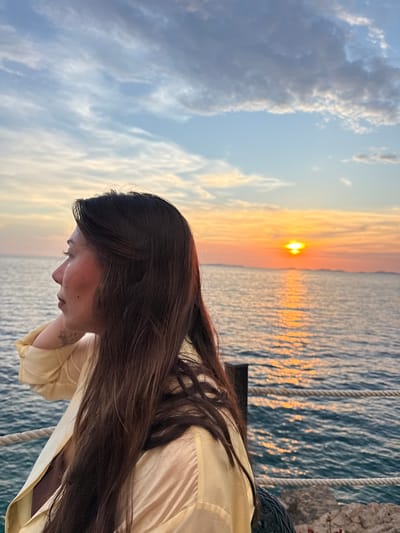 A side-profile medium shot captures a woman with long, dark hair looking out at the sea during sunset in Primošten, Croatia. She is wearing a pale yellow collared shirt, open to reveal a dark garment underneath. Her left hand is resting on the back of her neck, and a tattoo is visible on her arm. The background features the sea, a sunset with orange and yellow hues, and a hazy distant coastline. A rope railing is present in the foreground, and a dark post is visible to the right. The sky is partly cloudy with streaks of white and gray.