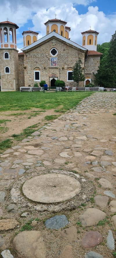 This image presents an exterior view of a stone church or monastery situated in Ribaritsa, Bulgaria, under a partly cloudy sky. The foreground is occupied by a pathway composed of irregular river stones or cobblestones set into a bed of gravel and dirt, with interspersed patches of green grass. A circular concrete cover is visible embedded in the path at the bottom-center of the frame.

In the midground, a grassy area with some bare earth extends towards the building. A series of dark-stained wooden benches are arranged in front of a white picket fence. A person in a light-colored top and shorts is seated on a bench on the right, alongside a dark-colored stroller. Another individual wearing a blue garment is partially visible near a green bench on the left. Trimmed green hedges are positioned directly in front of the church's facade.

The background features the stone church building, constructed from varied, irregular stone blocks. On the left, a prominent bell tower rises with two levels of arched window openings and a conical, tiled roof surmounted by a cross. The main structure is adorned with multiple domes, each featuring a white base, a yellow upper section, and topped with a cross. The central facade displays arched windows with white frames and a large, rectangular icon depicting religious figures, located above the main entrance. A small, circular window with a cross motif is positioned directly above the icon. Green trees and foliage are visible on the right side of the building.