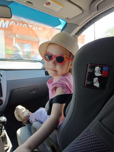 A young child is sitting in a car seat in the backseat of a car. The child is wearing a pink t-shirt, light purple pants, a beige straw hat, and sunglasses with red frames in the shape of hearts. The child is facing forward and looking to the left. The car seat is gray and black, with a label showing how to install the car seat for different weight classes. The child is wearing sneakers. In the background, a storefront with an orange facade and a sign displaying "MONETKA" in white letters can be seen. This suggests the location is near a Moneta store, likely in Ust-Kishert, Russia. The car is parked, and sunlight is coming in through the windshield. A gear shift is visible to the left of the child.