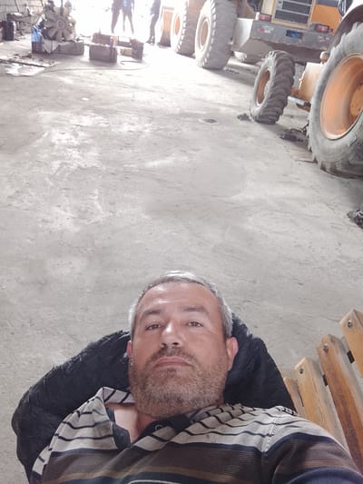 A man with a graying beard and striped shirt reclines in what appears to be a repair garage. Behind him, large machinery, possibly a bulldozer or similar heavy equipment, is parked. In the background, indistinct figures stand near what might be disassembled engine components. The exact location cannot be definitively determined to be Sabir Rakhimov, Uzbekistan, without further information.