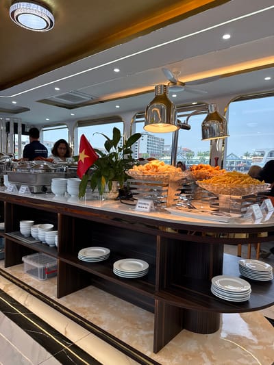 This image depicts a brightly lit interior of a buffet area, likely on a cruise ship or large boat, given the large windows and "HERCULES CRUISE" text on a uniform. The setting appears upscale with polished dark wood finishes, light-colored marble accents, and modern lighting fixtures including recessed spotlights, strip lights, and two prominent metallic industrial-style lamps illuminating the food. A small ceiling fan is also visible.

Through the curved windows, distant buildings are visible under what appears to be an overcast daytime sky, suggesting a waterfront or coastal location. This aligns with the provided context of Tuan Chau Ward, Vietnam.

The main activity is a buffet service. A long counter features several chafing dishes and clear serving platters holding various fried foods, including french fries and what looks like fried chicken or seafood. A small sign on the counter clearly labels one item as "SEAFOOD SPRING ROLLS" with its Vietnamese translation, "Nem hải sản chiên". Serving tongs are present for guests.

Two staff members are visible behind the buffet. One person with dark hair and glasses is attending to the food area, while another individual, partially seen, wears a dark uniform with "HERCULES CRUISE" printed on it. On the shelves adjacent to the buffet, stacks of clean white plates and bowls are neatly arranged for diners. A small Vietnamese flag is prominently displayed on the counter near a potted plant, further indicating the location.