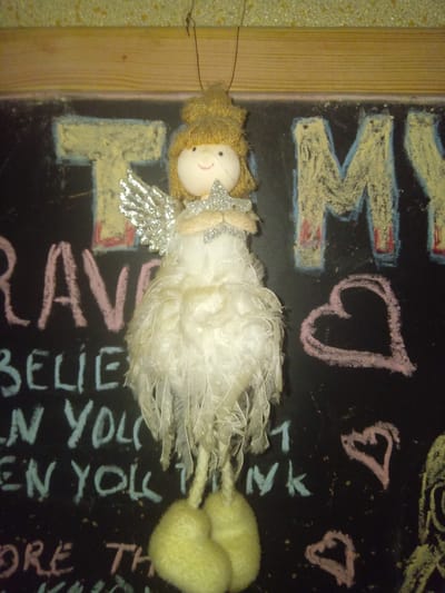 An overhead shot of a decorative angel ornament hanging in front of a chalkboard that has been drawn on with chalk. The angel is white with a yellow head and has a silver star in its hands. The chalkboard has the words "T" "MY", "RAVE", "BELIEVE", "IN YOU", "EN YOU", "nk", "ORE", "TH", and several heart drawings. The chalk writings appear to be motivational or inspirational quotes. The background of the image is out of focus but appears to be a wall with a yellow speckled texture. There is a wooden frame around the top and right sides of the chalkboard. The image was taken in Terebovlia, Ukraine.