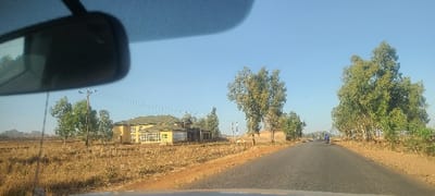 The image displays a daytime view of a paved road in Hoss, Nigeria, observed from inside a vehicle, with a rearview mirror partially visible at the top left. The sky is clear and blue. The asphalt road extends into the distance, bordered by dry, reddish-brown earth and sparse, yellowish vegetation. On the left side of the road, in the middle ground, a large, two-story structure with a light yellow facade and a dark, possibly tiled, roof is visible. This building features multiple windows, including a curved section on its left. A wooden utility pole with overhead lines stands near the building. Directly opposite, on the right side of the road, a white and black striped pole or sign is erected. Numerous trees with green foliage are distributed along both sides of the road and in the background. In the far distance along the road, a single individual is operating a blue motorcycle, moving away from the camera. Distant hills or rock formations are discernable on the horizon to the far left.