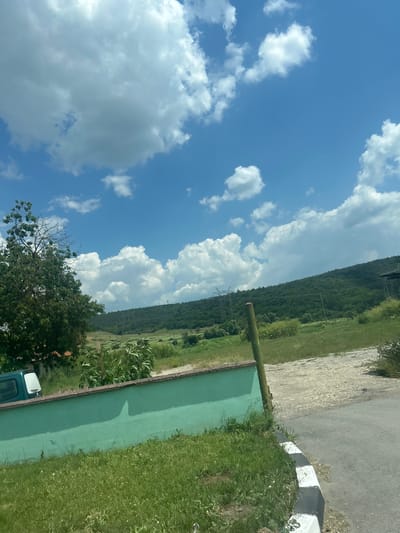 In General Kiselovo, Bulgaria, this image captures a rural landscape under a bright, blue sky dotted with fluffy white clouds. The foreground features a low, bright green wall topped with a concrete border, bordering an area of lush green grass. To the left, the corner of a dark green vehicle is visible, parked behind the wall. Beyond the wall, the terrain slopes upwards, covered in green vegetation and trees, leading to a densely forested hill in the background. Power lines and transmission towers are visible on the slopes of the hill, indicating the presence of infrastructure. A dirt path runs alongside the green wall, and a paved road curves into the lower right of the frame, marked with black and white striped concrete curbs. The overall scene conveys a sense of calm, natural beauty with subtle signs of human presence.