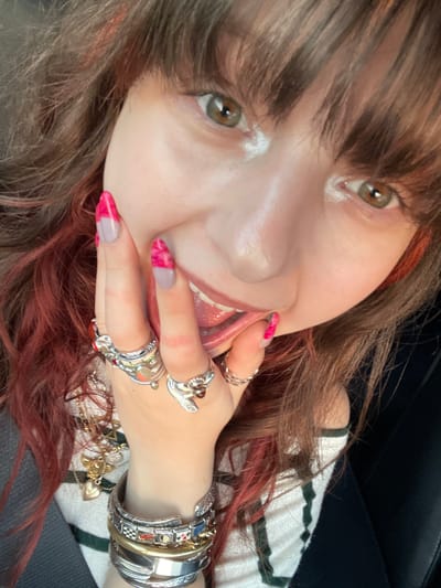 The image is a close-up selfie of a woman. She has brown hair with red highlights and light brown eyes. Her hand is near her face and mouth, with her tongue partially visible. She has several rings on her fingers and bracelets on her wrist. She appears to be wearing a white and green striped shirt. The background shows a dark surface. There is no information to indicate that this image was taken in the city of Hod HaSharon, Israel.