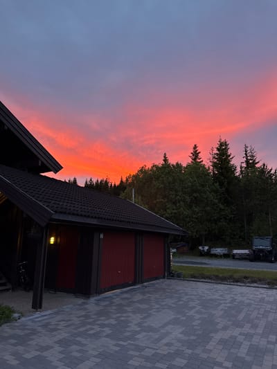 The image depicts a residential setting at dusk in Åmot, Norway. The sky is a prominent feature, displaying a vibrant gradient of colors ranging from deep blue at the top to fiery red and orange near the horizon, indicative of a sunset. In the foreground, a dark-tiled roof of a house or garage is visible, with a section of red garage doors. A light fixture is illuminated on the side of the structure. Adjacent to the building, a paved driveway composed of grey and black interlocking stones leads away from the viewer. A bicycle is parked on the gravel area near the entrance. Beyond the house, a dense line of dark green coniferous trees forms the background, silhouetted against the colorful sky. In the distance, on the right side of the frame, several trailers and a dark-colored all-terrain vehicle are parked on a gravel area. The environment suggests a rural or semi-rural location.