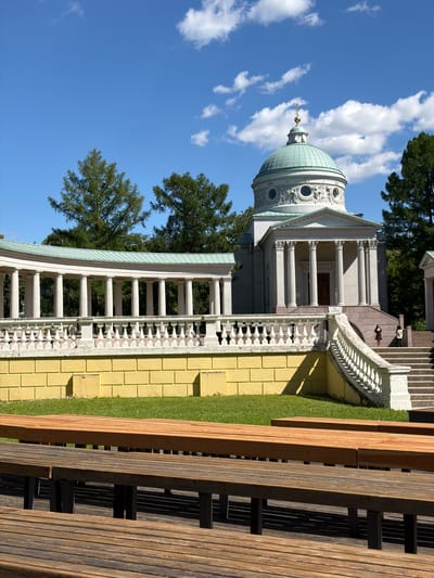 The image shows a neoclassical building with a dome, possibly a chapel or mausoleum, located in the estate of Arkhangel'skoye, Russia. The building is white with a teal dome topped with a golden cross. It features a grand portico with tall columns and a raised entrance approached by a wide staircase. A long, curved colonnade with similar columns extends from the left of the building, also topped with a teal roof. The structures are set against a backdrop of lush green trees and a bright blue sky with scattered white clouds. In the foreground, there are several rows of empty wooden benches, suggesting an outdoor seating area or performance space. Two individuals are visible on the steps of the main building, one standing at the bottom of the stairs and another further up. The architecture and landscaping indicate a historical and potentially significant site.
