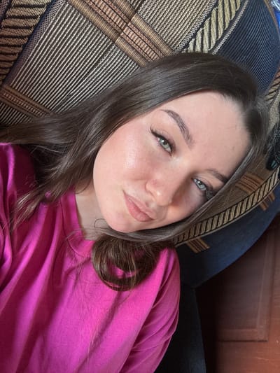 The media file is a close-up selfie of a young woman lying on a patterned couch. She has long, dark brown hair and is wearing a vibrant pink shirt. Her head is tilted, and she is looking towards the camera with a slight smile and her lips pursed. Her eyes, which appear to be green or hazel, are looking directly at the viewer. Her eyebrows are well-defined, and she has noticeable eyeliner and mascara on her lashes.
The background shows the textured fabric of a couch with a striped pattern in shades of brown, beige, and black. A dark blue cushion is visible behind her. The lighting suggests it is daytime, possibly indoors, with soft light illuminating her face. The overall mood is relaxed and casual. The image is cropped to focus on the woman's face and upper body, with the couch filling the lower portion of the frame. There is no visible text in the image. The location context provided is Krasnokamsk, Russia.