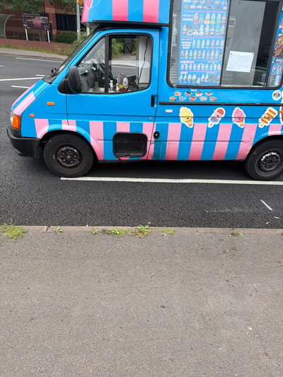 An ice cream van, painted bright blue with pink stripes, is parked on an asphalt road with a white line marking the parking space. The van has a menu displayed in its window, featuring various ice cream options like cones, sundaes, and trays. Stickers of ice cream characters adorn the side of the van. The surrounding environment includes a paved curb with some weeds growing, and a building with advertising visible in the background. The scene is captured from a side-view, at approximately eye-level.