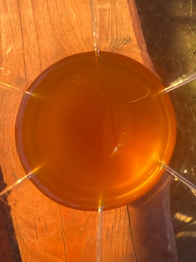 This is a top-down, close-up shot of a clear glass filled with an amber-colored liquid, likely a beverage. The glass is positioned on a wooden surface that appears to be weathered and textured, with visible wood grain and slight cracks. Sunlight is hitting the scene, creating bright highlights and casting a warm glow, suggesting it is daytime. The liquid has a smooth surface with subtle ripples and a few small, light-colored specks floating within it. The edges of the glass are visible, extending upwards and inwards, with reflections of the surrounding environment, including what appears to be vegetation and possibly the sky, visible on the glass. The overall impression is one of a simple, still-life scene, possibly outdoors given the lighting and natural elements.