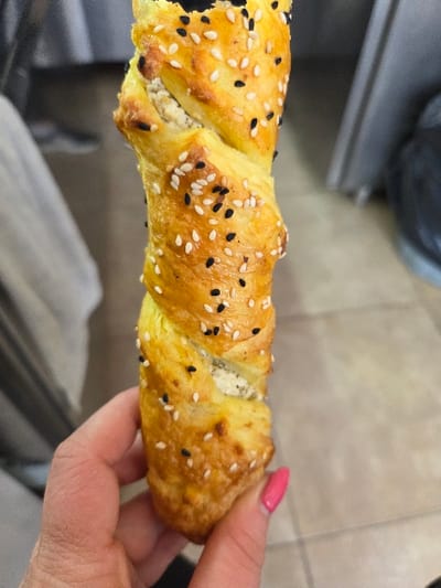 A close-up, eye-level shot depicts a person's hand holding a single, golden-brown, twisted pastry, situated indoors in the city of Gevgelija, North Macedonia. The pastry is cylindrical, covered with white sesame seeds and black nigella seeds, and shows a light-colored, possibly cheese, filling exposed along its twisted form. The hand has a fair complexion, with the thumb and forefinger supporting the pastry from below. The visible fingernail on the forefinger is painted with pink polish. The background is out of focus, revealing a blurred light-colored fabric on the left and a darker, possibly gray surface with a light-colored tiled floor visible on the right.
