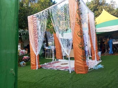 An outdoor event scene in Amokvo, Nigeria, features a central, open-sided canopy structure with four primary support columns draped in pleated, shimmering orange-brown fabric. Green artificial vine garlands are affixed to some columns. White, translucent fringe-like curtains hang vertically from the canopy's frame, enclosing the space. Beneath the canopy, a circular rug with a central pattern and a red, white, and black floral border rests on artificial green turf that covers the entire visible ground.
To the left of the canopy, an ornate, high-backed chair with a white cushion and golden accents is visible, surrounded by clusters of artificial flowers in pink, purple, white, and blue hues. Further left, a solid green vertical surface partially frames the image.
To the right and rear of the canopy, a large tent with a prominent yellow conical roof section and horizontal green and white stripes provides shelter. Underneath this tent, a person in a light blue jumpsuit operates multiple electronic keyboards or music production equipment set up on a stand. Another individual is partially visible near the musician. Lush green foliage from trees forms the background.