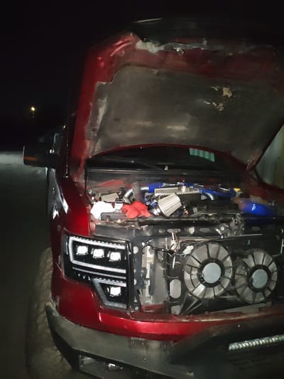 A red pickup truck is parked at night, with its hood open, revealing its engine and cooling fans. The truck has custom headlights with multiple square LED lights. The scene appears to be outdoors, possibly in a rural or industrial area, given the dark surroundings and the presence of a streetlamp in the background. There are no people visible in the image. The truck is the sole subject of the photograph. The location is Krtsanisi, Georgia.