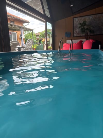 The image depicts an indoor swimming pool with vibrant turquoise water, reflecting the light and creating rippling patterns on the surface. The pool is framed by a glass enclosure, allowing a view of a backyard with a house, lounge chairs, and greenery. In the background, inside the enclosure, are several large, bright pink beanbag chairs and a piece of art on the wall. Two metal pool ladders are visible, partially submerged in the water. The scene suggests a resort or a private residence designed for relaxation and recreation. The location is identified as Chiprovtsi, Bulgaria.