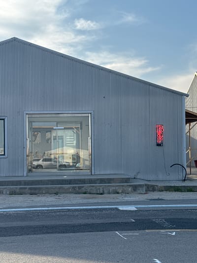 The image shows the exterior of a building in Toseong-myeon, South Korea. The building is constructed of corrugated metal and has a steeply pitched roof. A large window dominates the facade and reflects the interior of the building, showing what appears to be display cases and objects. A neon sign is mounted above the window. A sidewalk runs along the base of the building, with a street visible on the right side of the frame. Markings are on the road. The sky is partially cloudy.