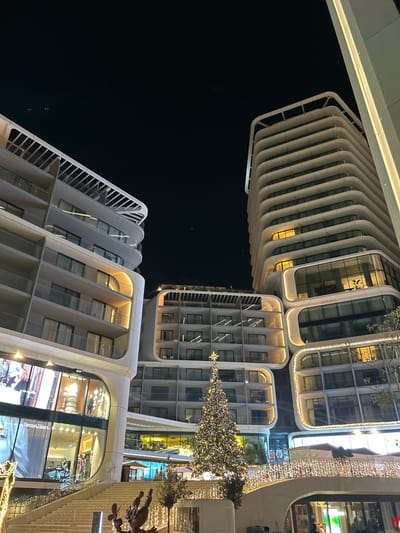 Modern architecture dominates the night scene. Arched, illuminated buildings frame a dark sky. A Christmas tree glittering with lights stands between structures. One building features stacked, rounded segments, while another showcases a more angular design with numerous windows. Architectural lighting enhances the unique shapes and edges of the buildings, creating a contemporary urban landscape.