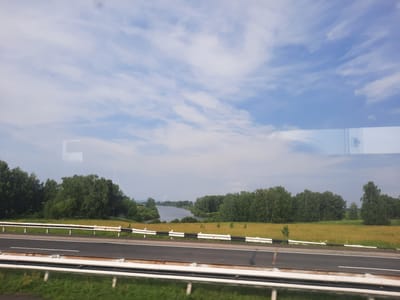 The image, taken from a moving vehicle, shows a highway in Smirnovskii, Russia. The road is asphalt with white lane markings. A white guardrail with black and white reflective strips separates the highway from a field of dry, yellow grass. Beyond the field, a dense line of green trees forms the background, with a body of water, likely a river, visible through a gap in the trees. The sky above is partly cloudy, with streaks of white clouds against a pale blue background. The scene is natural and rural, with no people or buildings visible.